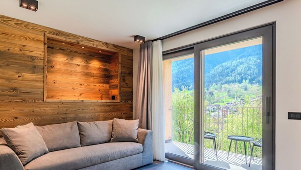 Un salon moderne avec un canapé, un mur recouvert de panneaux en bois et une porte-fenêtre coulissante donnant sur un balcon avec vue sur un paysage verdoyant.