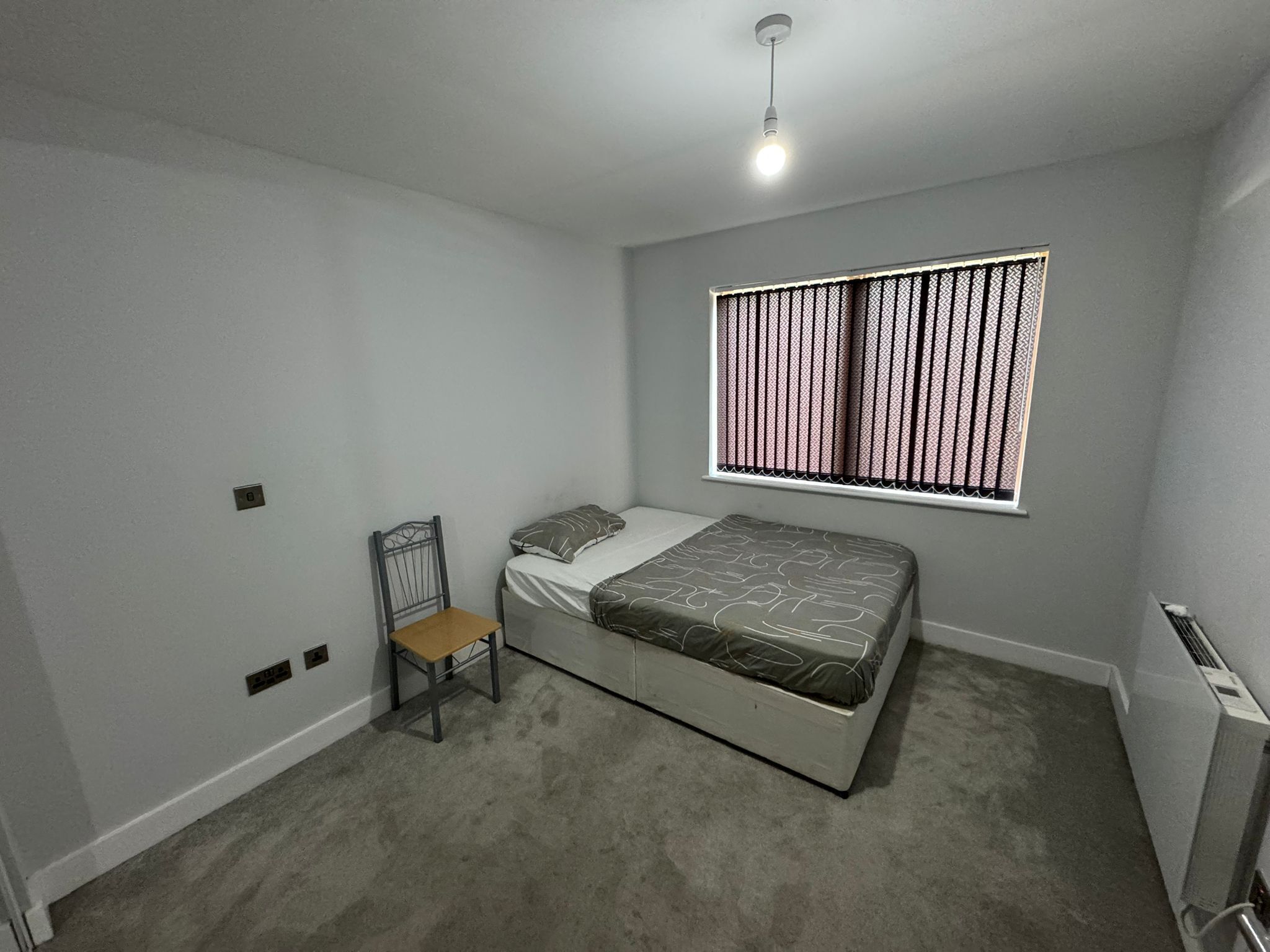 Un dormitorio con cama, una silla, una ventana con persianas y una lámpara en el techo.