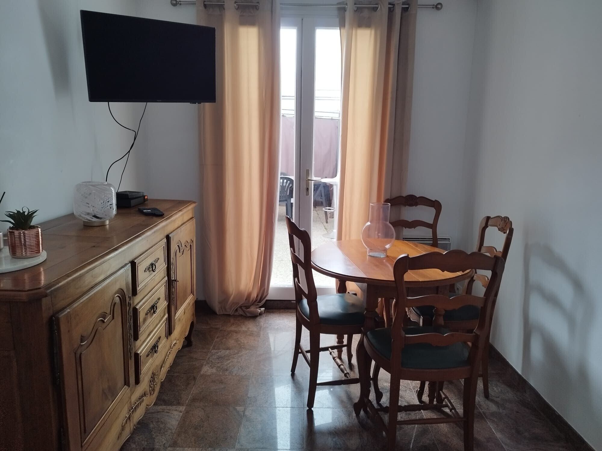 A dining area with a wooden table and chairs, a television mounted on the wall, and a window with curtains.