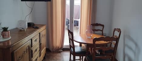 A dining area with a wooden table and chairs, a television mounted on the wall, and a window with curtains.