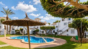 A swimming pool with a thatched umbrella, surrounded by well-maintained grass and palm trees, with white buildings in the background. - Playa Flamenca With Terrace (Orihuela)