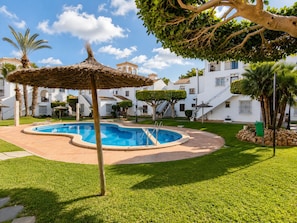 A swimming pool with a thatched umbrella, surrounded by well-maintained grass and palm trees, with white buildings in the background. - Playa Flamenca With Terrace (Orihuela)