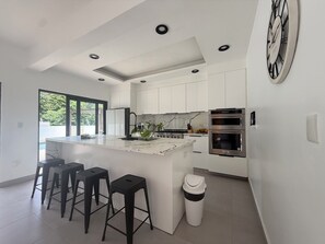 Una cocina moderna con armarios blancos, una isla de mármol, taburetes negros, un cubo de basura y hornos integrados.