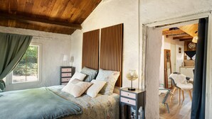 Une chambre avec un plafond en bois, un lit recouvert d’une literie à motifs floraux, une table de chevet avec une lampe, et une vue sur un coin cuisine.