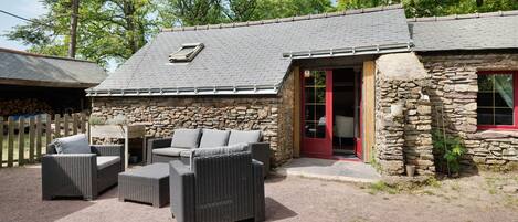 A stone building with a grey tiled roof, a red door, and wicker furniture outside.