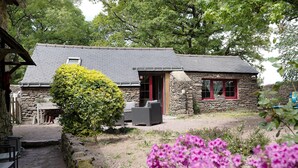 Une charmante maison en pierre avec une porte rouge, un espace extérieur aménagé pour s’asseoir et un jardin parsemé de fleurs roses.