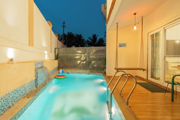 A swimming pool with a waterfall feature, a metal ladder, and a glass door leading to an outdoor area. - Alaya Stays Cavalo Marinho 3 BHK (Colvale)