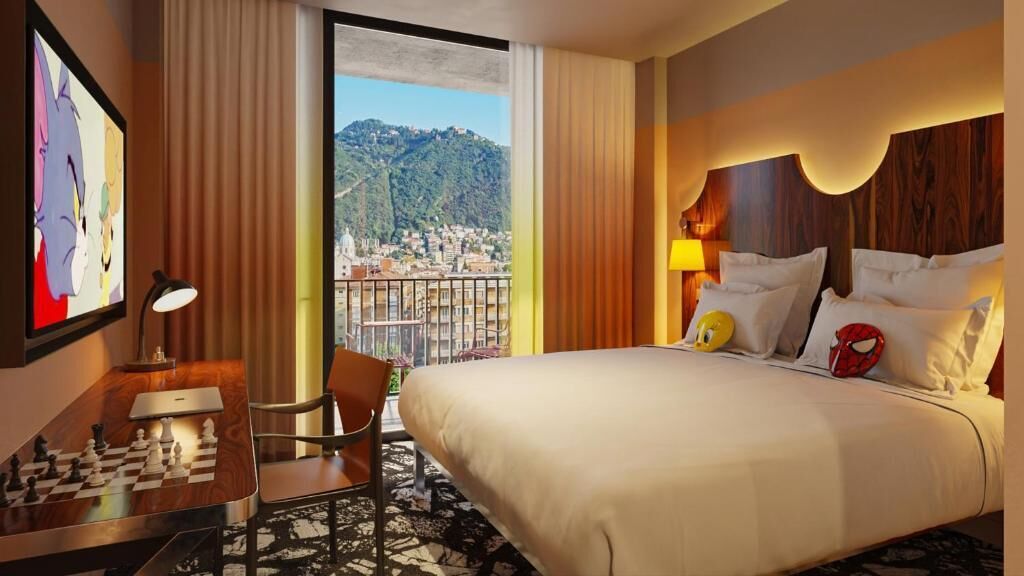 Une chambre d’hôtel avec un lit, un bureau sur lequel se trouve un jeu d’échecs, une chaise, une lampe et une grande fenêtre donnant sur la ville et les collines.