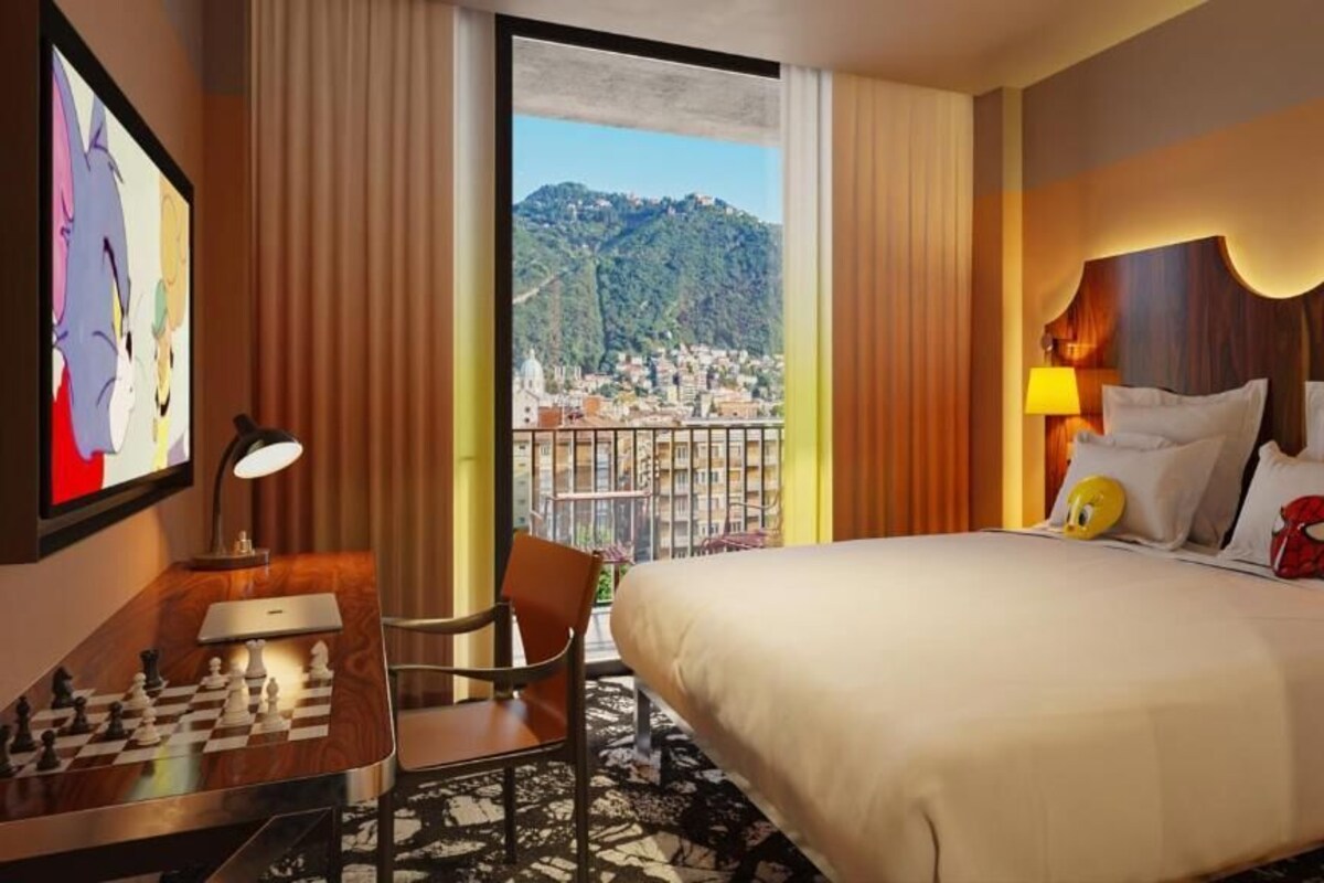 A hotel room with a bed, a desk with a chessboard, a chair, a lamp, and a large window with a view of a city and hills.