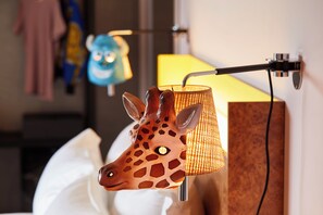 A giraffe-shaped lamp with a woven shade, a blue plush toy, and a yellow lamp in a room with a white wall and a wooden panel. - Mama Shelter Lake Como  (Como)