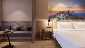 A hotel room with a bed, a sofa, a small table with fruit, a nightstand with a lamp, and a wall mural of a mountain landscape.