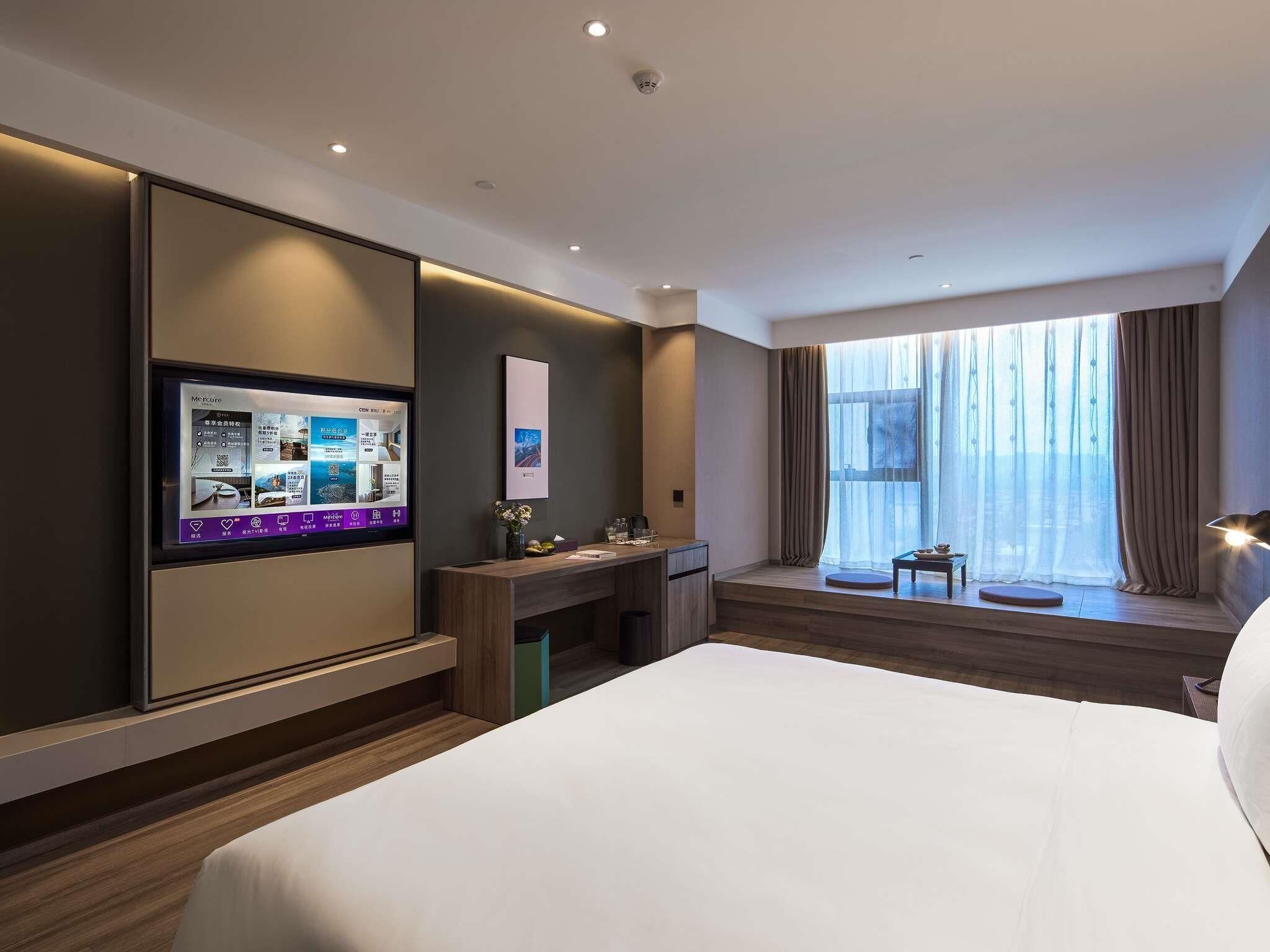 A hotel room with a bed, a desk, a TV, a window with curtains, and a view of the city.