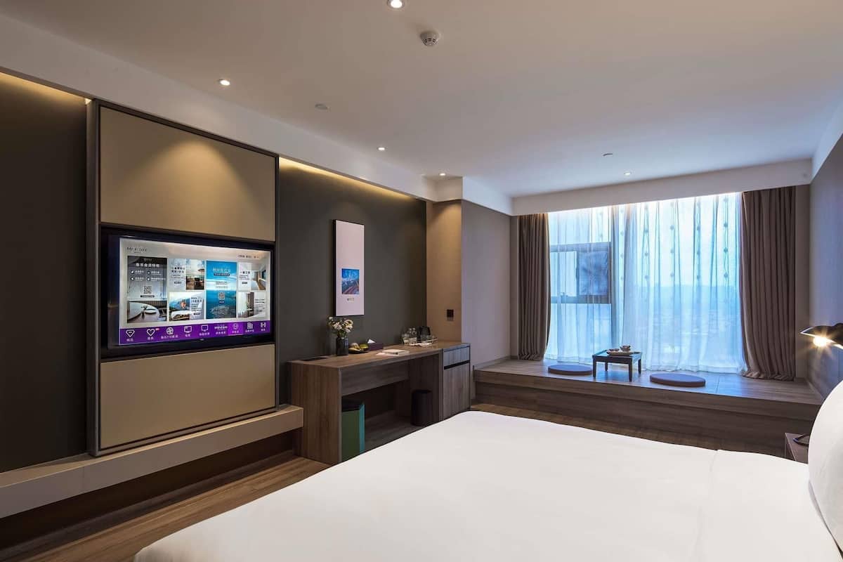 A hotel room with a bed, a desk, a TV, a window with curtains, and a view of the city.