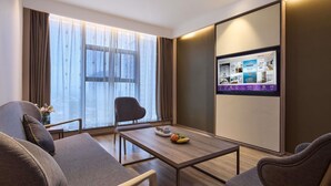 A modern hotel room with a sofa, a dining table, a TV mounted on the wall, and a large window with curtains.