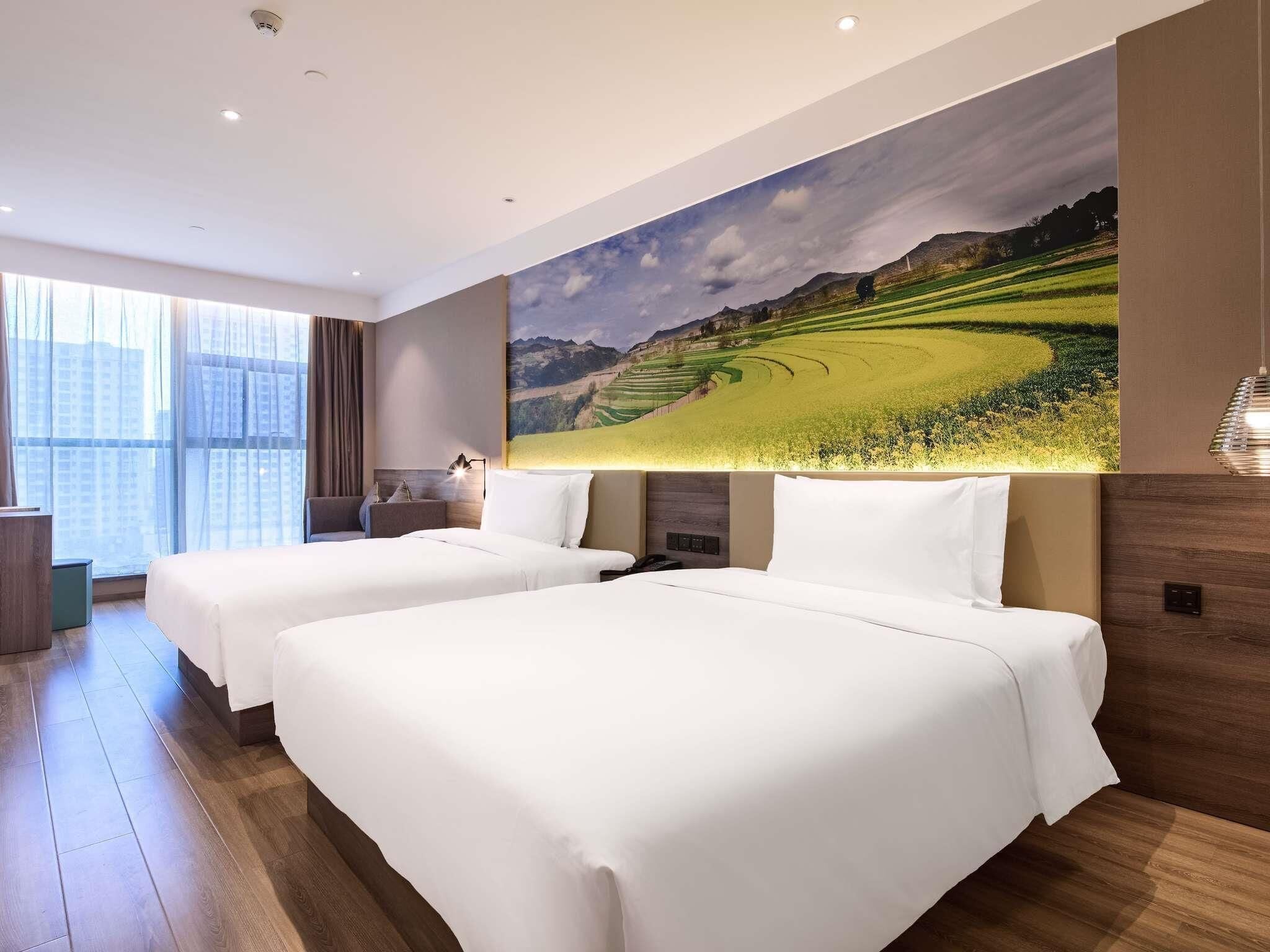 A hotel room with two beds, a large window, and a wall-mounted painting of a landscape.