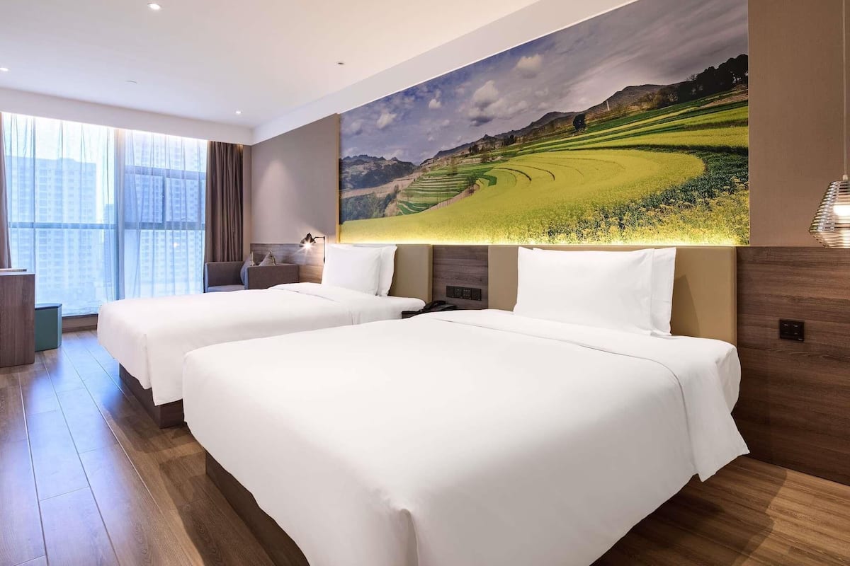 A hotel room with two beds, a large window, and a wall-mounted painting of a landscape.