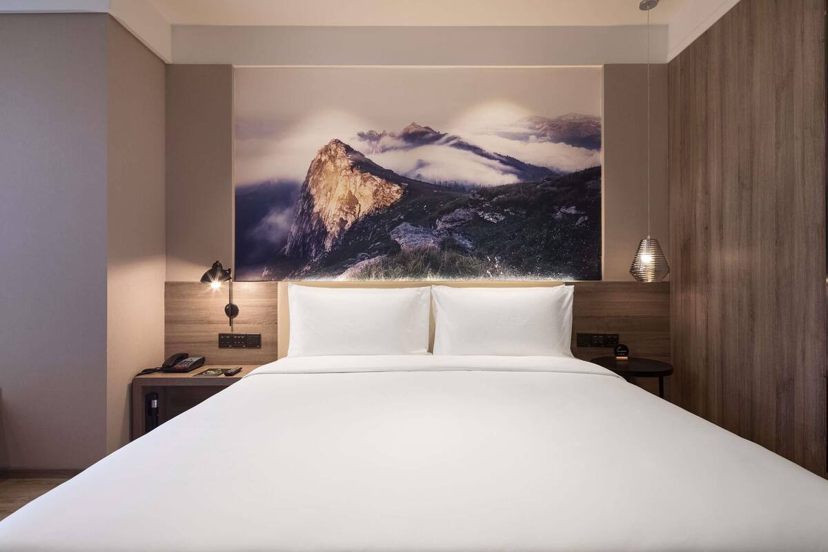 A modern hotel room with a large bed, a mountain landscape mural, bedside lamps, and a wooden accent wall.
