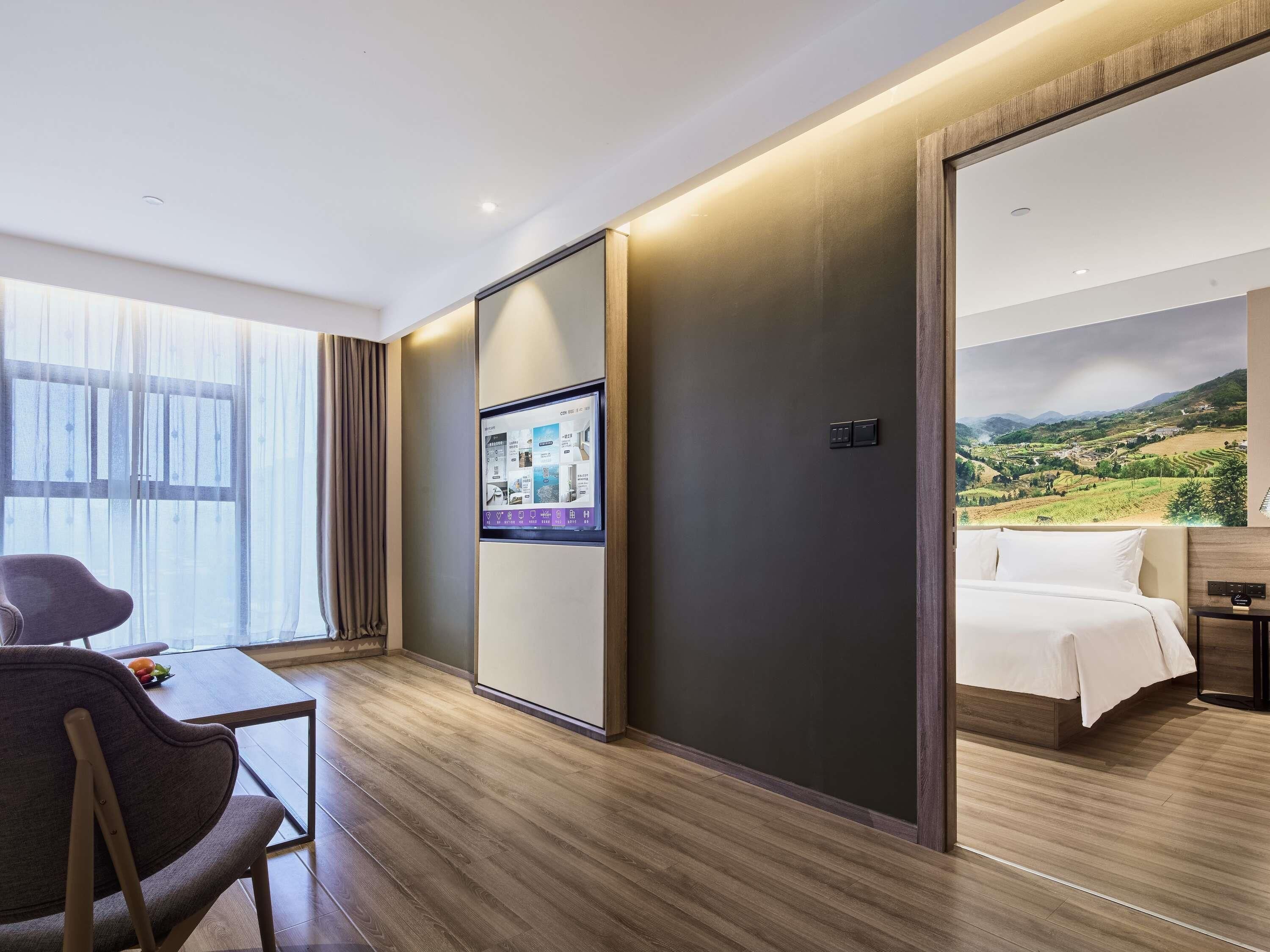 A hotel room with a bed, a desk with a chair, a TV screen, and a large window with curtains.
