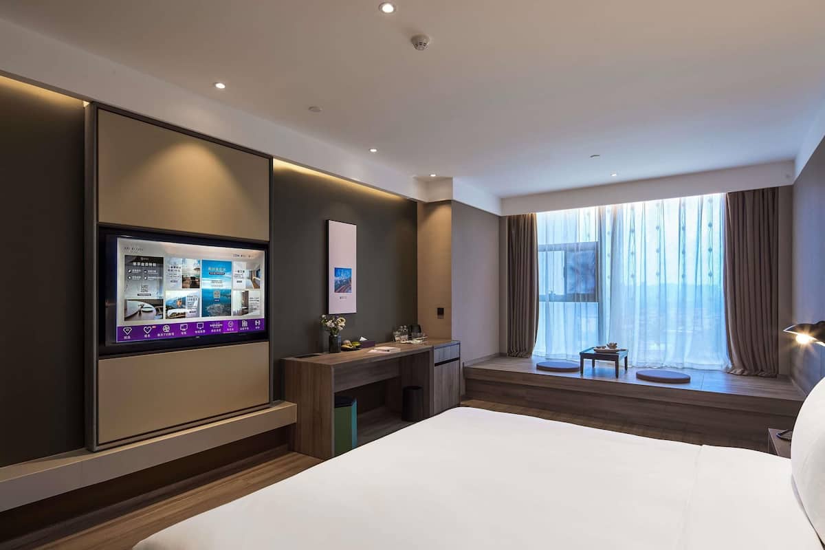 A hotel room with a large bed, a desk with a computer, a window with curtains, and a view of the city.