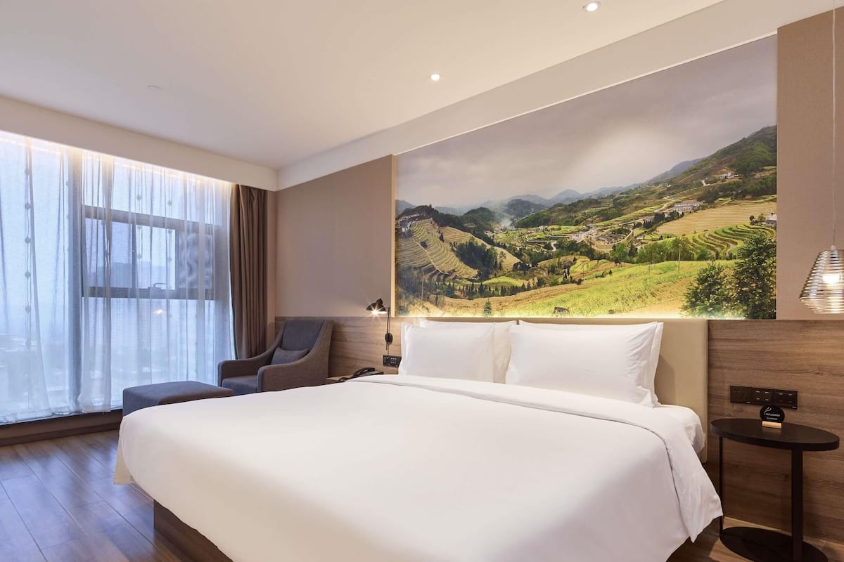 A hotel room with a large bed, a chair, a bedside table, a lamp, and a large landscape painting on the wall.