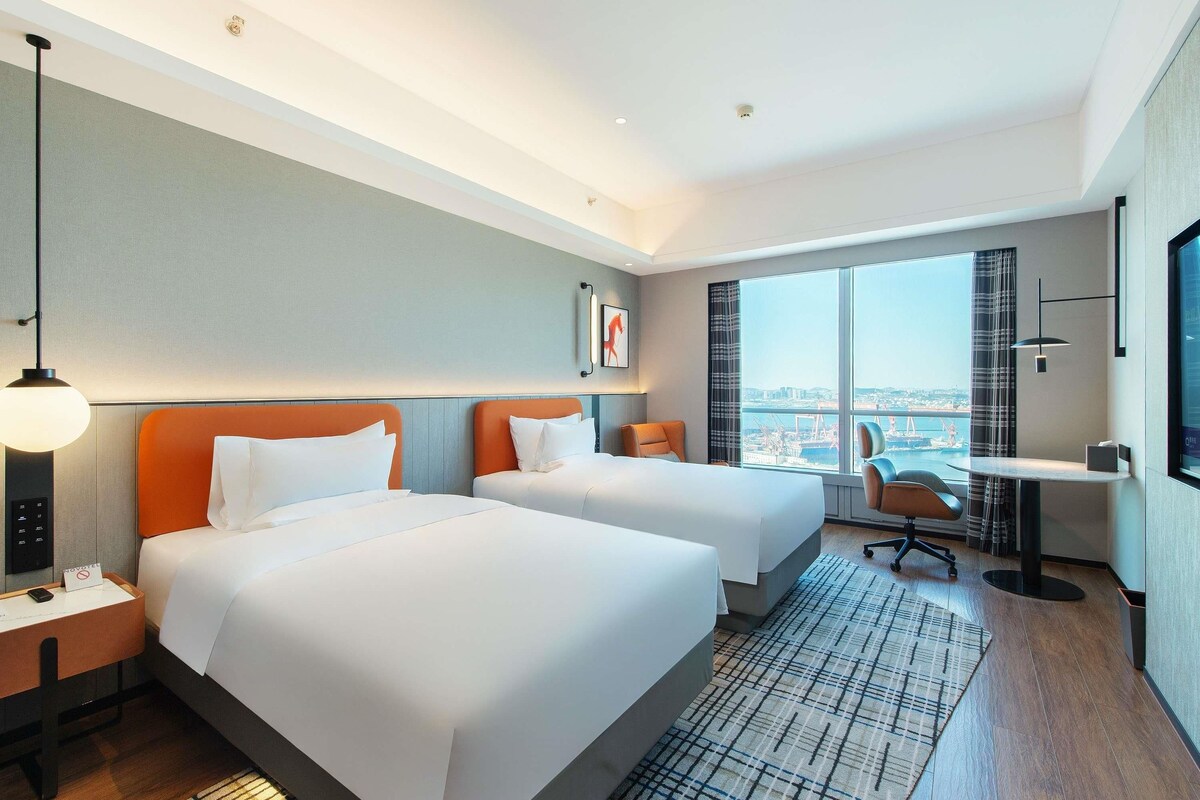 A hotel room with two beds, a desk, a chair, a TV, and a large window with a city view.
