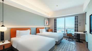 Habitación de hotel con dos camas, un escritorio, una silla, un televisor y un ventanal con vista a la ciudad.
