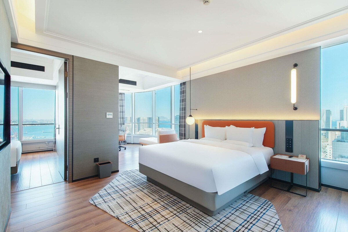 Ein modernes Hotelzimmer mit einem großen Bett, Blick auf die Stadt, einem Schreibtisch und einem Sessel.