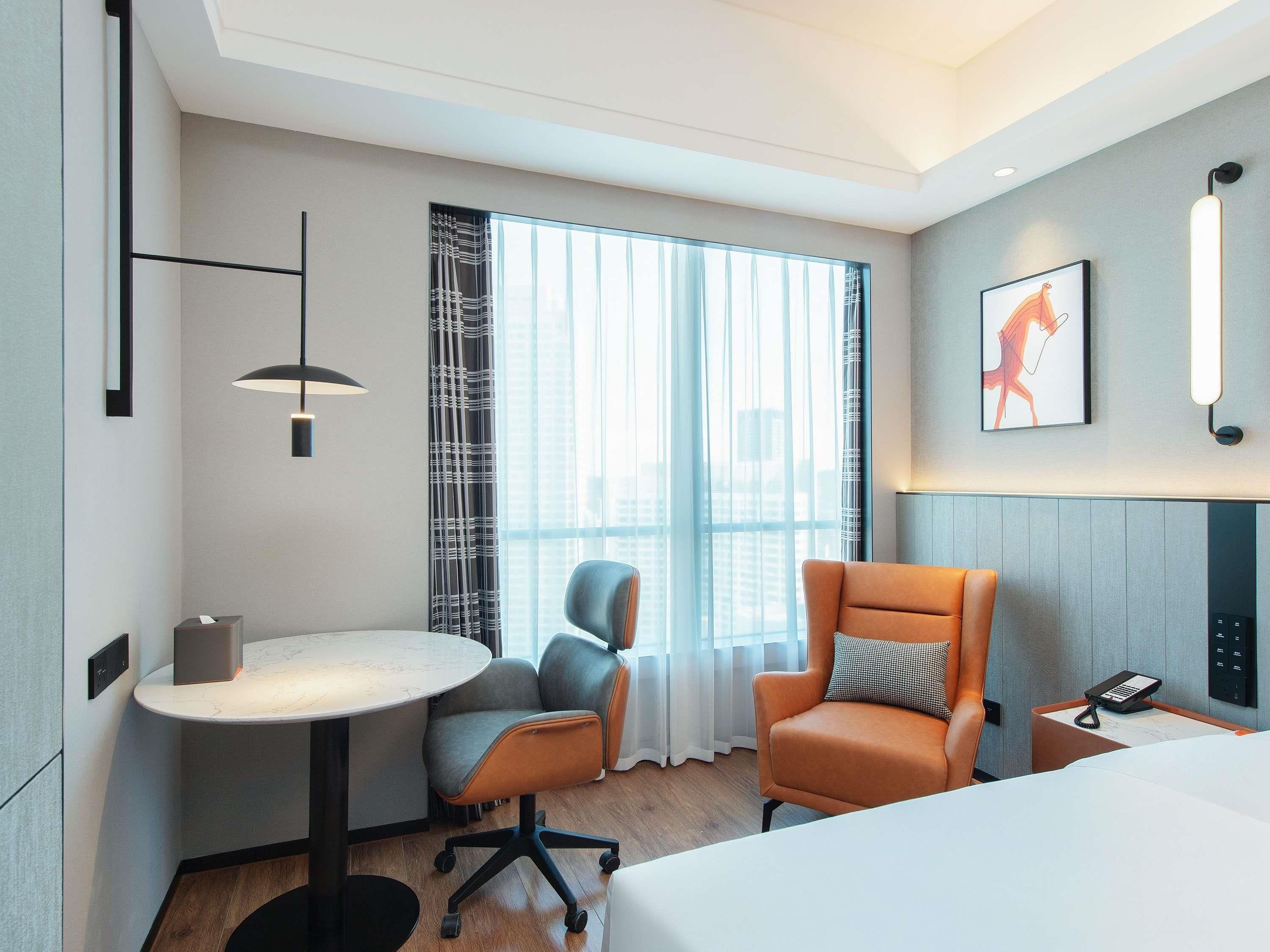 Habitació d'hotel amb un llit, una taula de treball amb una cadira, un telèfon sobre una taula auxiliar, una finestra amb cortines i un lluminària penjada a la paret.