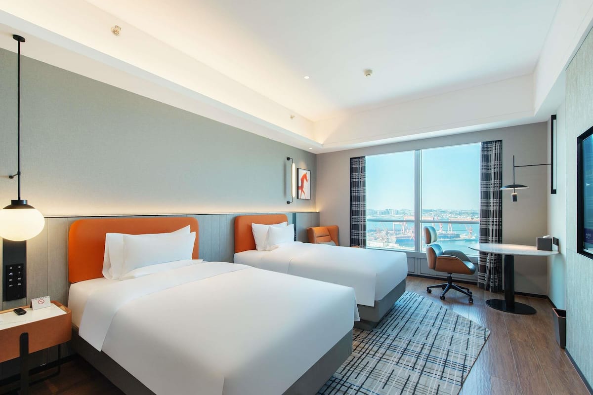 Habitación de hotel con dos camas, un escritorio, una silla, un televisor y un ventanal con vista a la ciudad.
