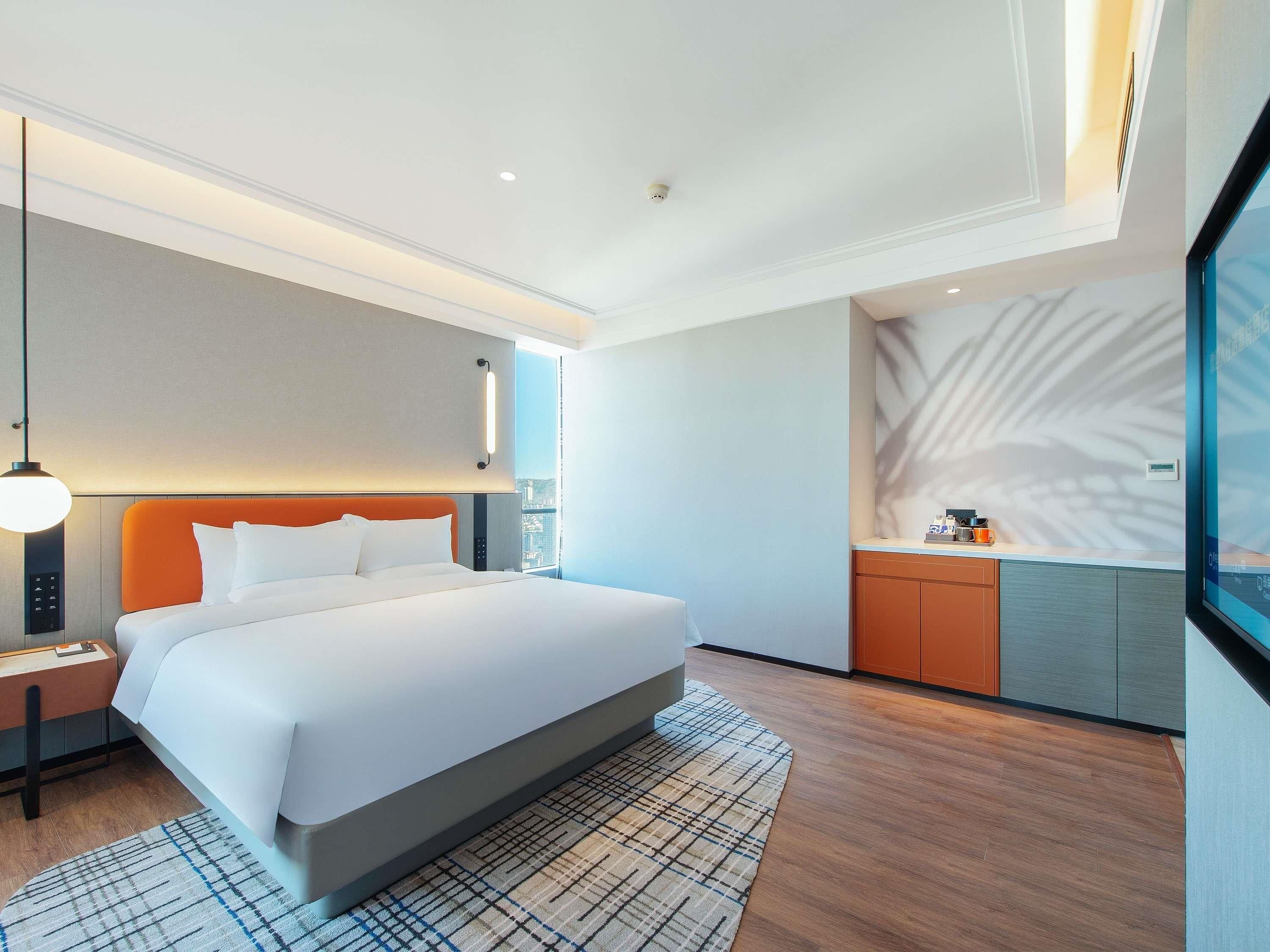 A modern hotel room with a large bed, a nightstand, a desk, and a view of the city.