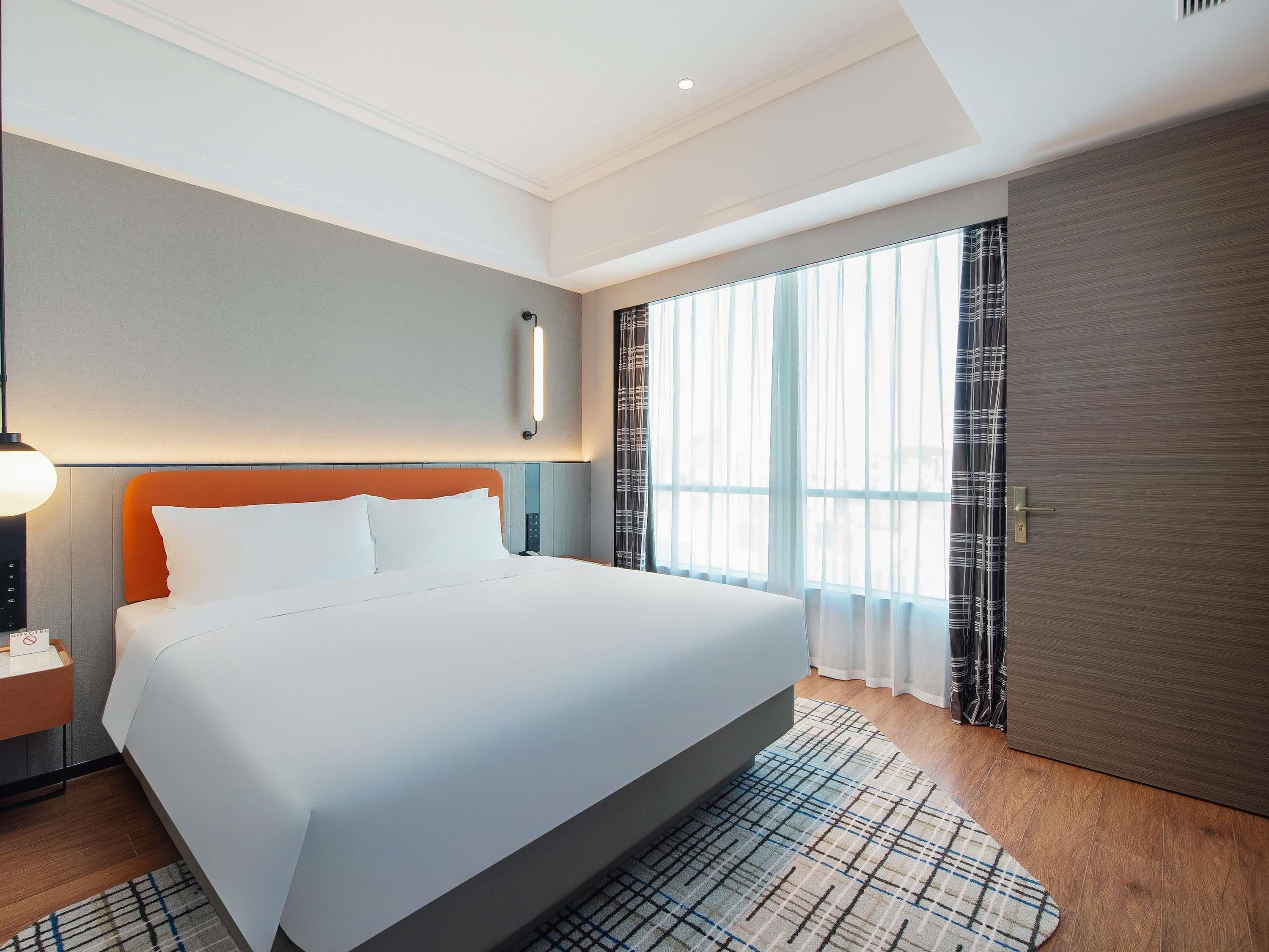 Una habitación de hotel con una cama grande, una mesita de noche con lámpara, una ventana con cortinas y una puerta.
