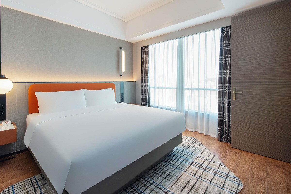 A hotel room with a large bed, a bedside table with a lamp, a window with curtains, and a door.