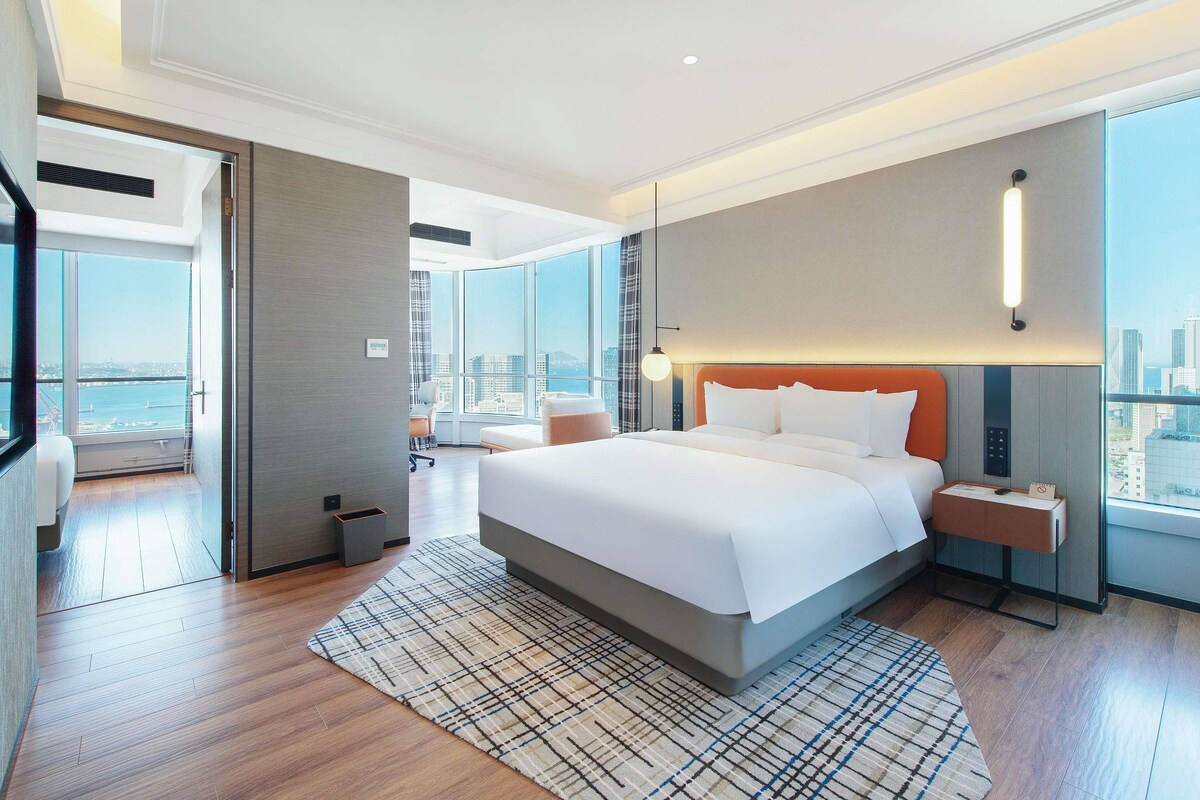 A modern hotel room with a large bed, a city view, a desk, and a chair.