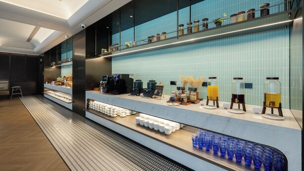 Un hall d’hôtel moderne avec un long comptoir d’accueil, un espace dédié au café et un mur recouvert de carreaux où sont disposés des pots.