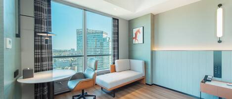 A modern hotel room with a large window, a desk, a sofa, and a wall-mounted phone.
