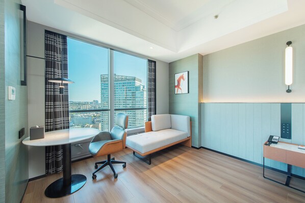 A modern hotel room with a large window, a desk, a sofa, and a wall-mounted phone.