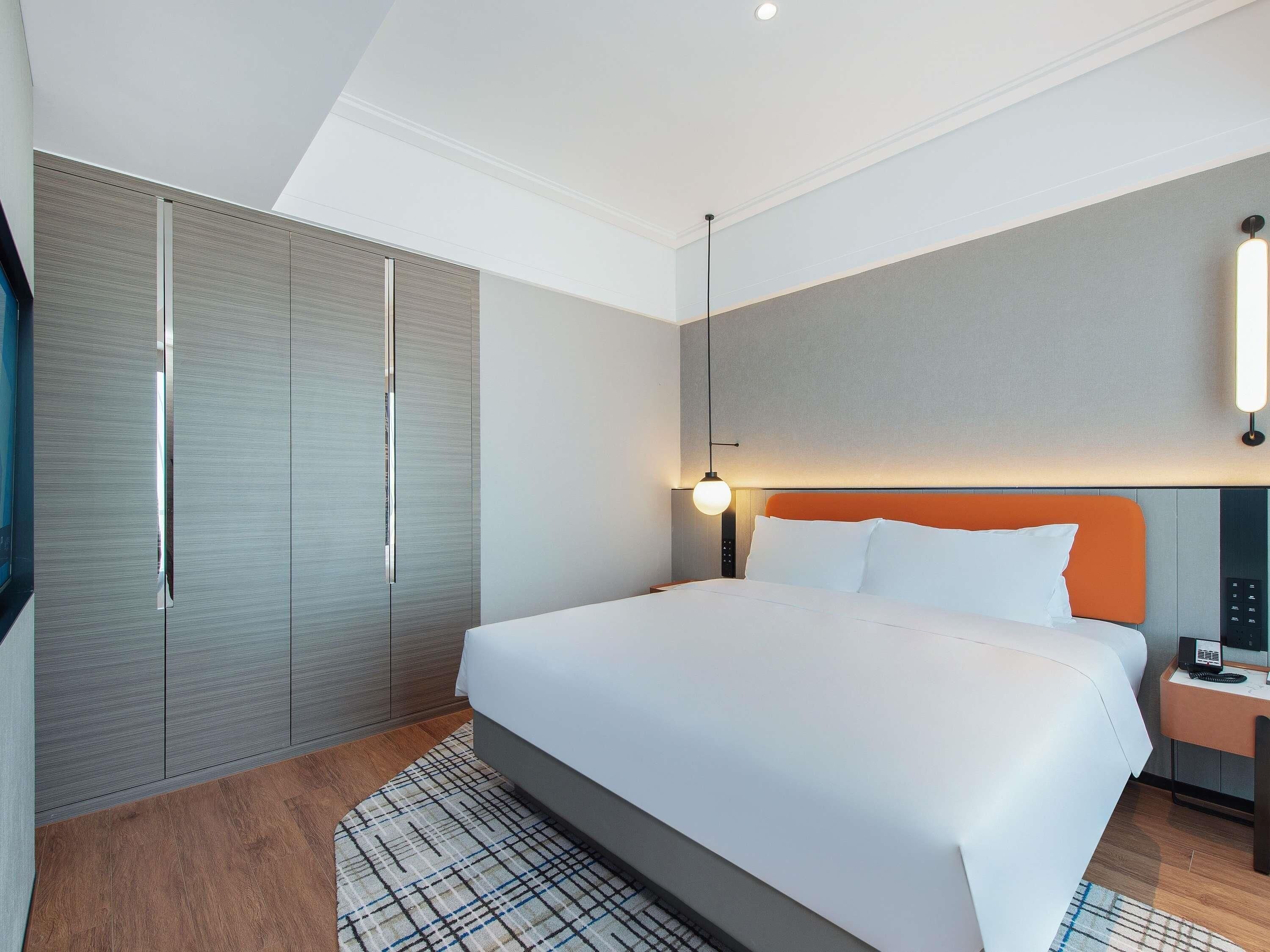 A modern hotel room with a large bed, a bedside table with a phone, a wardrobe, and a TV.
