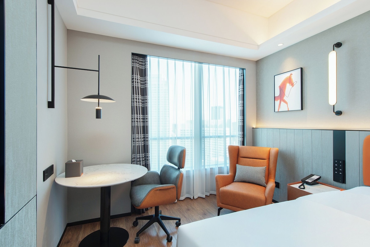Ein Hotelzimmer mit Bett, Schreibtisch mit Stuhl, einem kleinen Tisch, Telefon und einem Fenster mit Vorhängen.