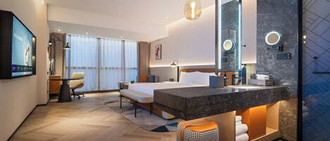 A modern hotel room with a large bed, a desk, a chair, a TV, a bathroom with a mirror, and a large window.