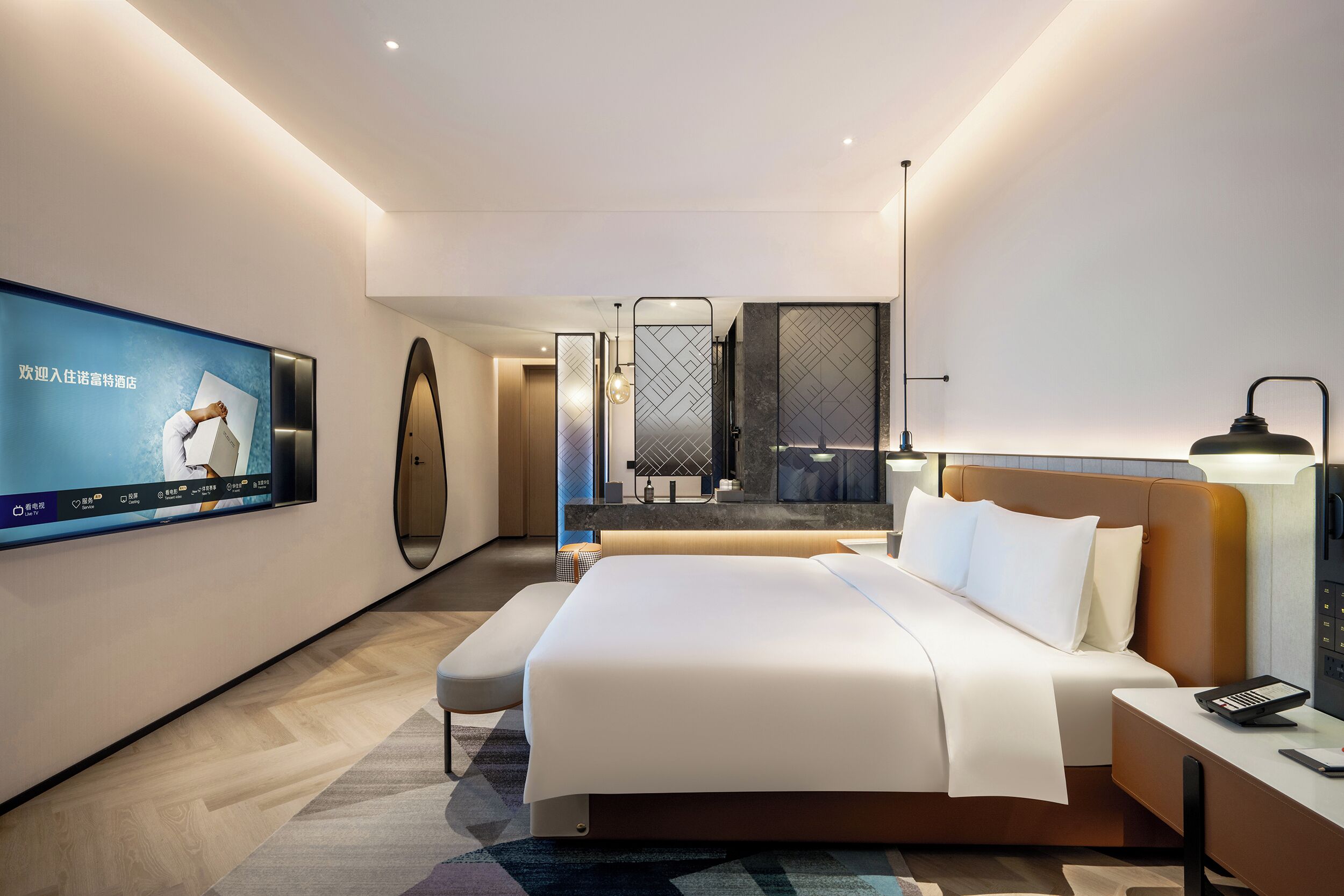 Habitación de hotel moderna con una cama grande, una mesita de noche con lámpara, un televisor montado en la pared y un baño visible a través de una mampara de vidrio.