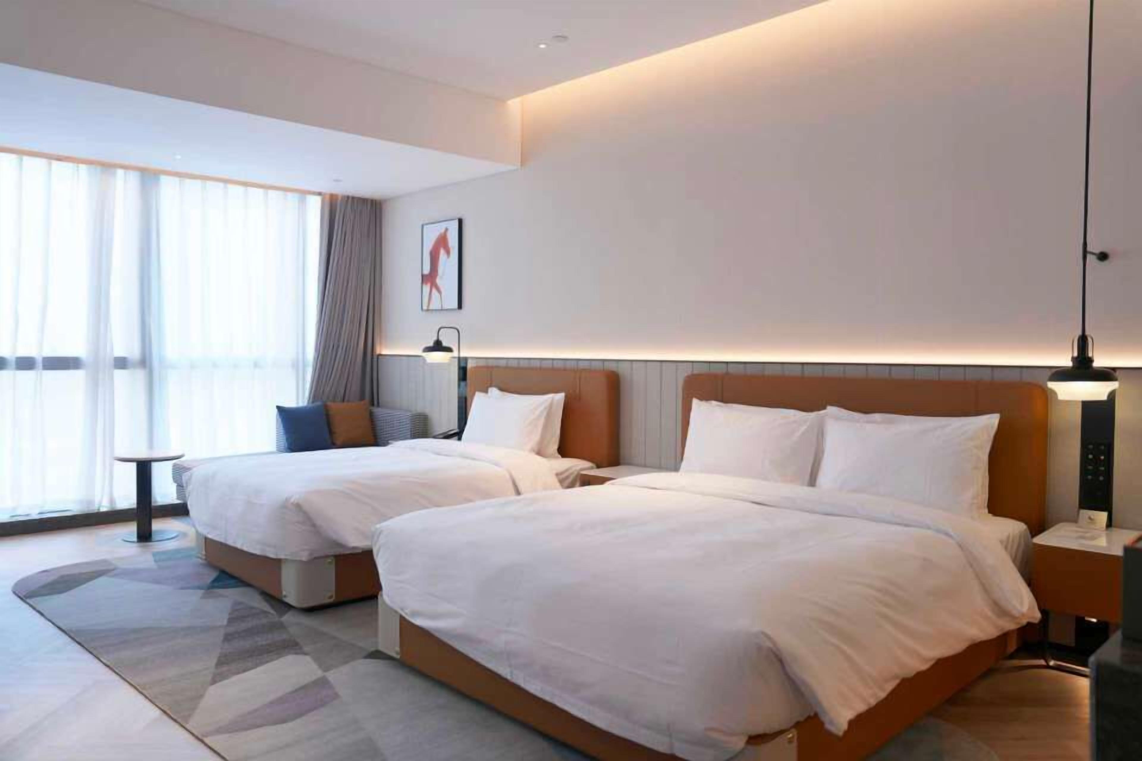 Une chambre d’hôtel avec deux lits, une petite table, une lampe et une grande fenêtre avec des rideaux.