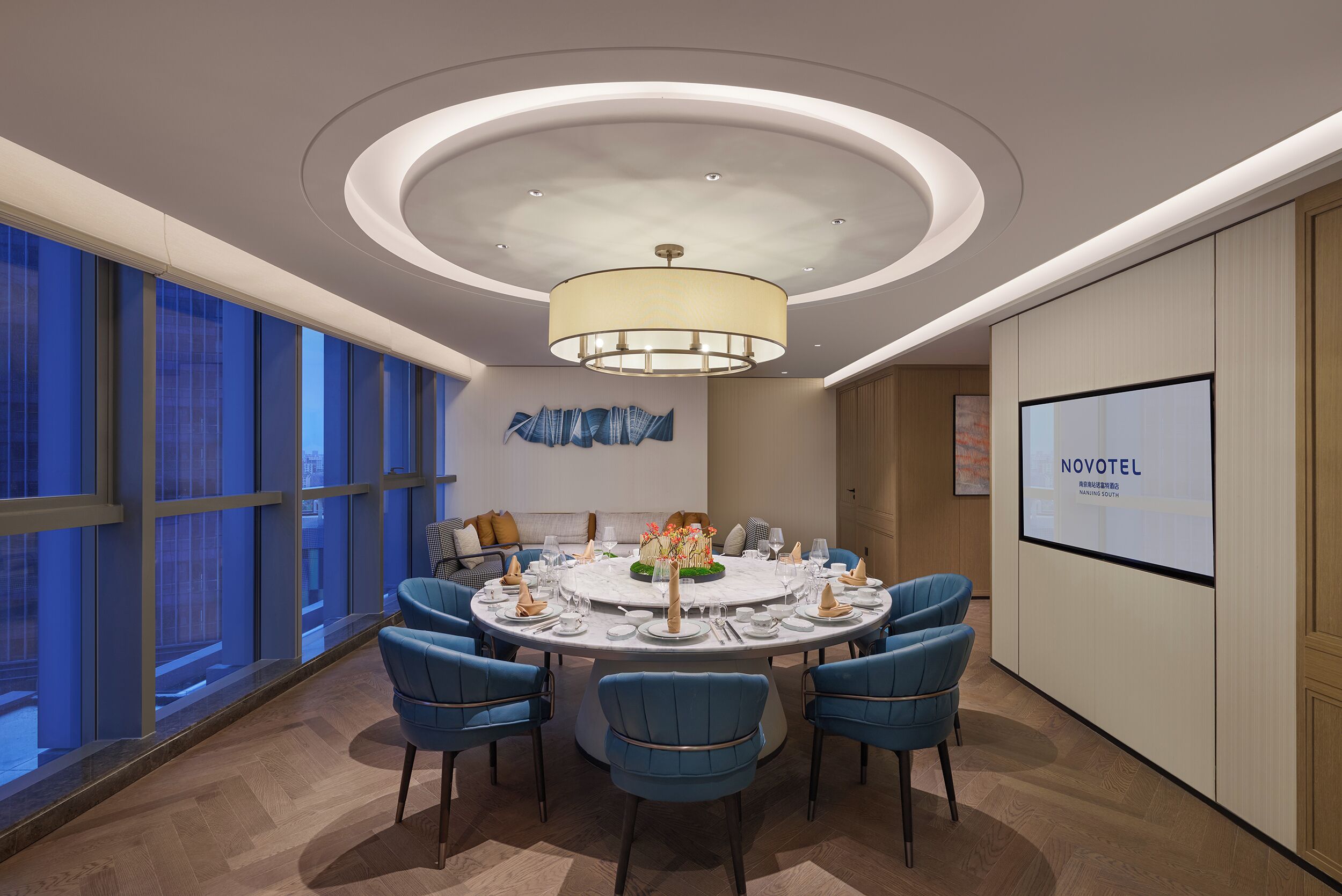 A modern hotel room with a round dining table, blue chairs, a large window, and a ceiling light fixture.