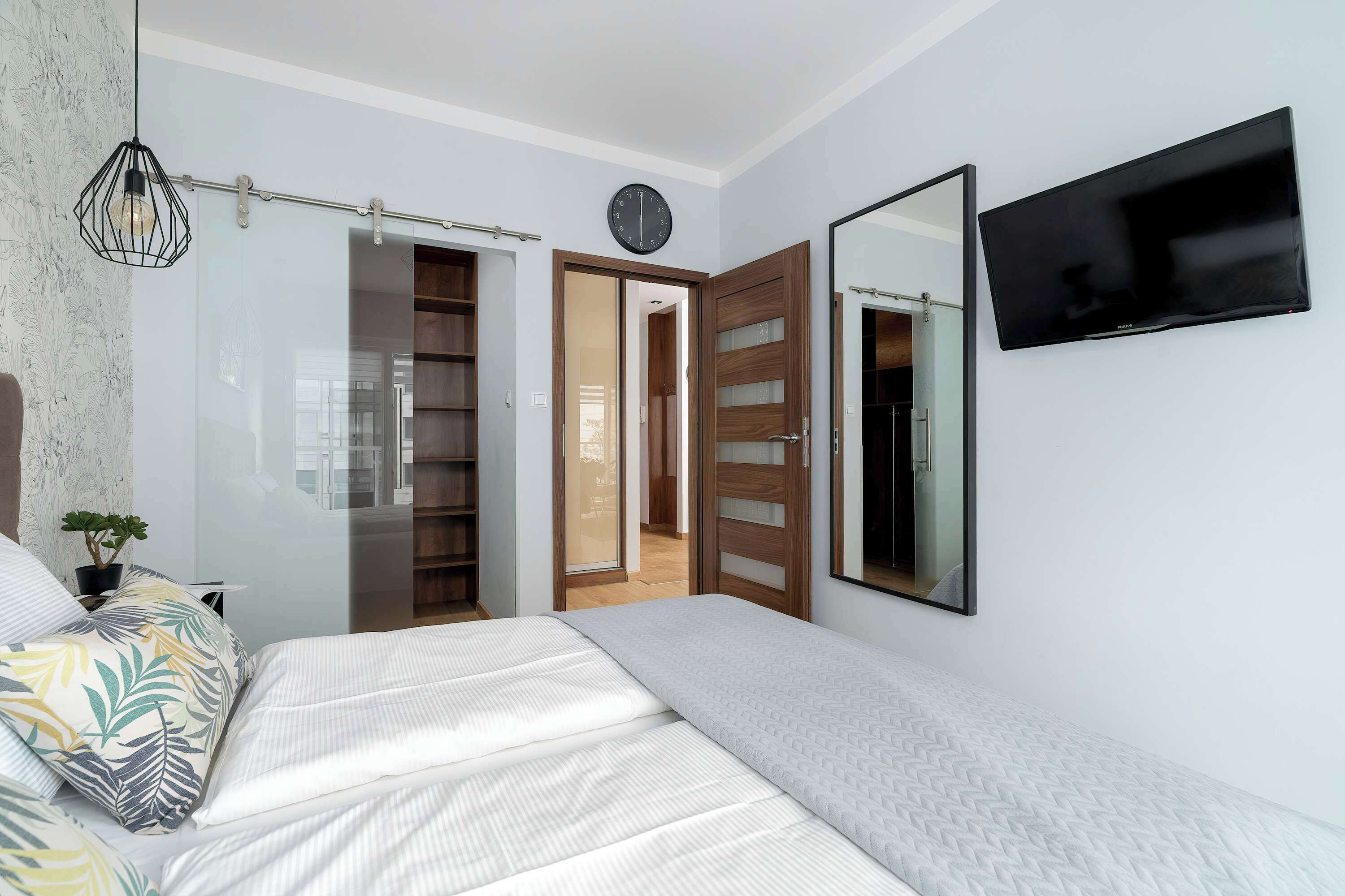 A bedroom with a bed, a bedside table with a plant, a wall-mounted TV, a clock, and a sliding door leading to a bathroom.