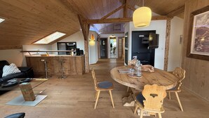 A cozy loft-style living space with a wooden ceiling, a dining table with chairs, a kitchen area, and a black leather sofa. - Vacation apartment Haus Bader (Garmisch-Partenkirchen)