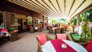 Een terras met rieten meubels, tafels met rood-witte tafelkleden en een overdekt gedeelte met houten plafond.