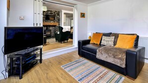 Ein Wohnzimmer mit einem Flachbildfernseher, einem schwarzen Ledersofa mit orangen Kissen, einem gemusterten Teppich und einem Kamin aus Stein.