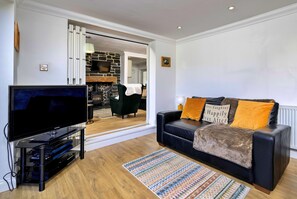 A living room with a flat-screen TV, a black leather sofa with orange cushions, a patterned rug, and a stone fireplace. - Dyffryn (Betws-Y-Coed)