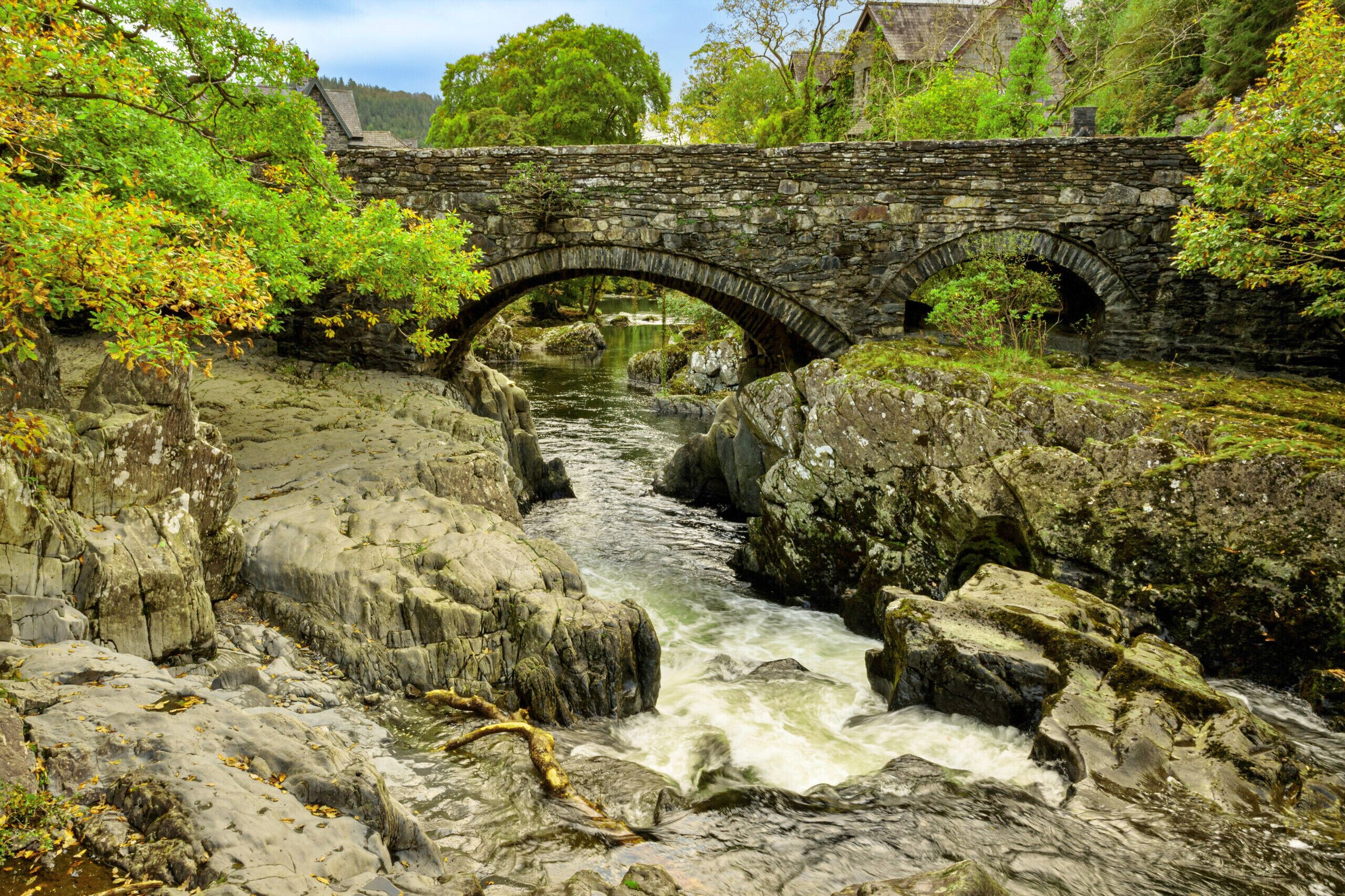 A stone bridge over a river with surrounding greenery and a building in the background.