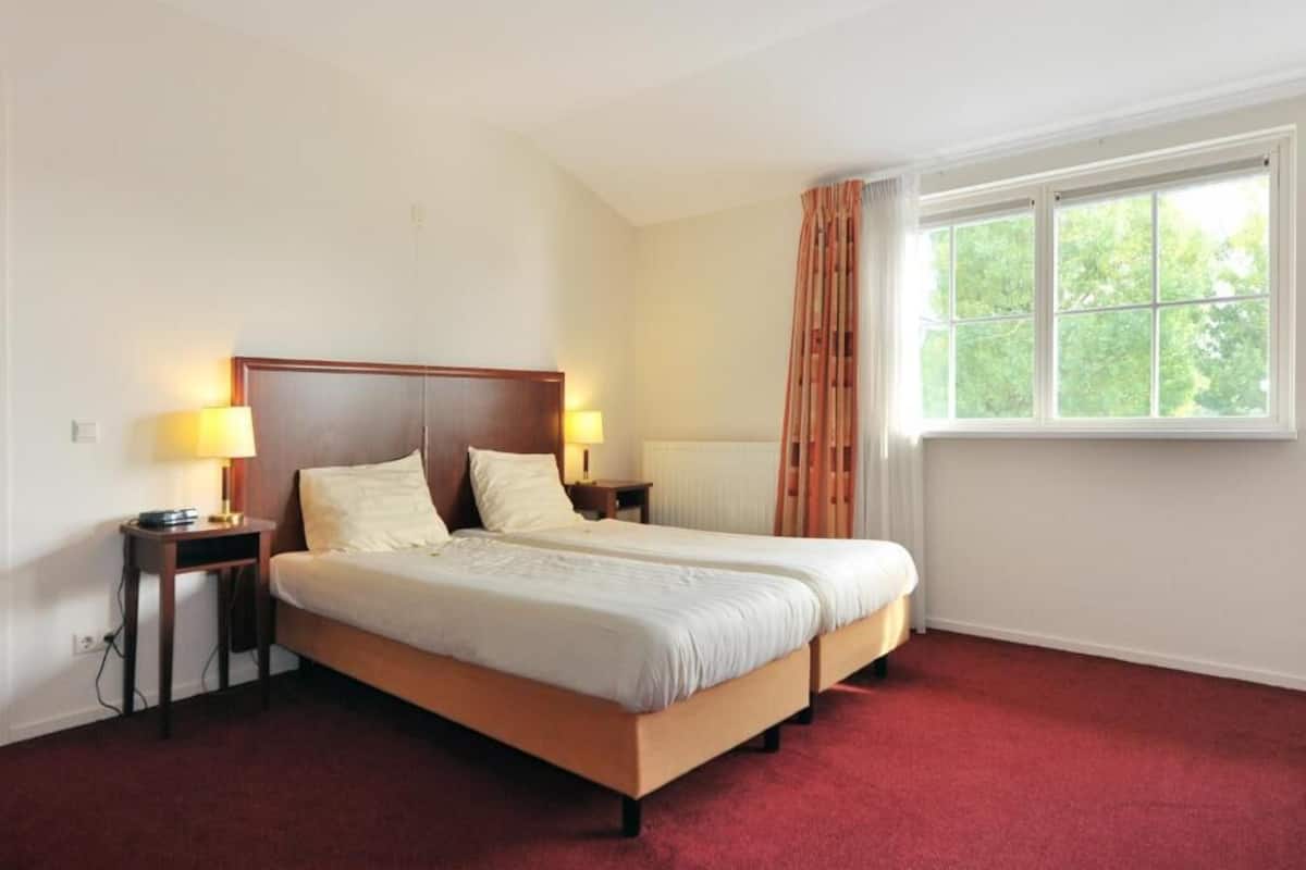 A double bed with headboards, two bedside tables with lamps, a window with curtains, and a red carpeted floor.