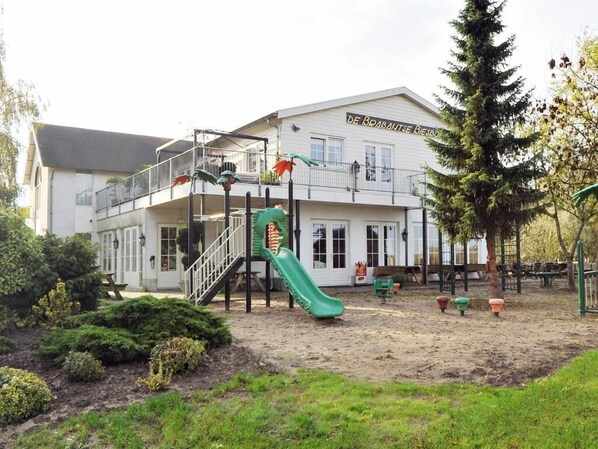 A building with a playground, including a slide and climbing structures, surrounded by greenery and a sandy area.